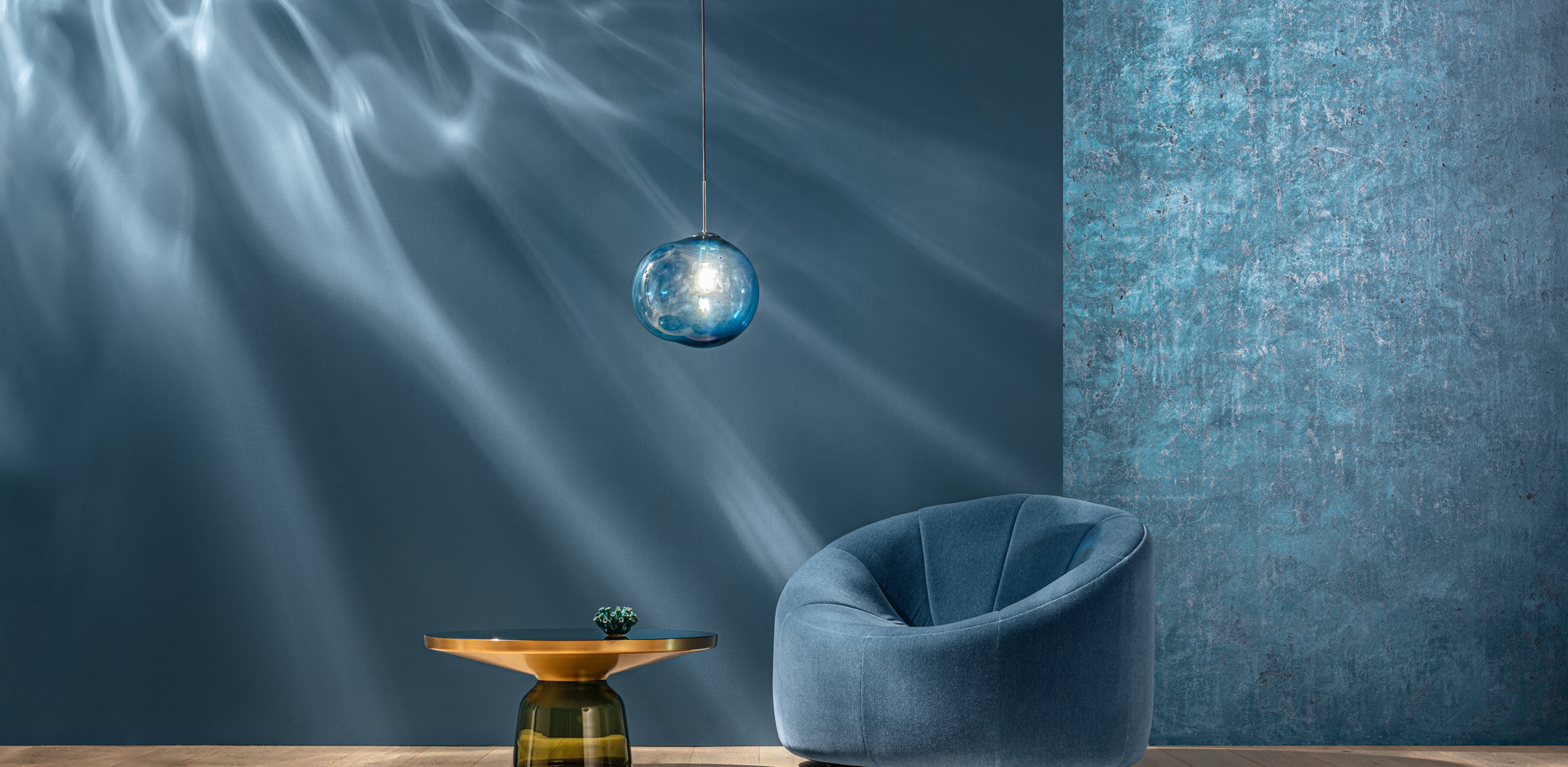 Ein stilvoll eingerichteter Raum mit einer runden blauen Couch und einem eleganten runden Tisch. Über dem Tisch hängt eine durchsichtige Lampe, die sanftes Licht ausstrahlt. Die Wände sind in dunklen Blautönen gehalten und zeigen eine sanfte Wellenbewegung, die an Wasser erinnert.